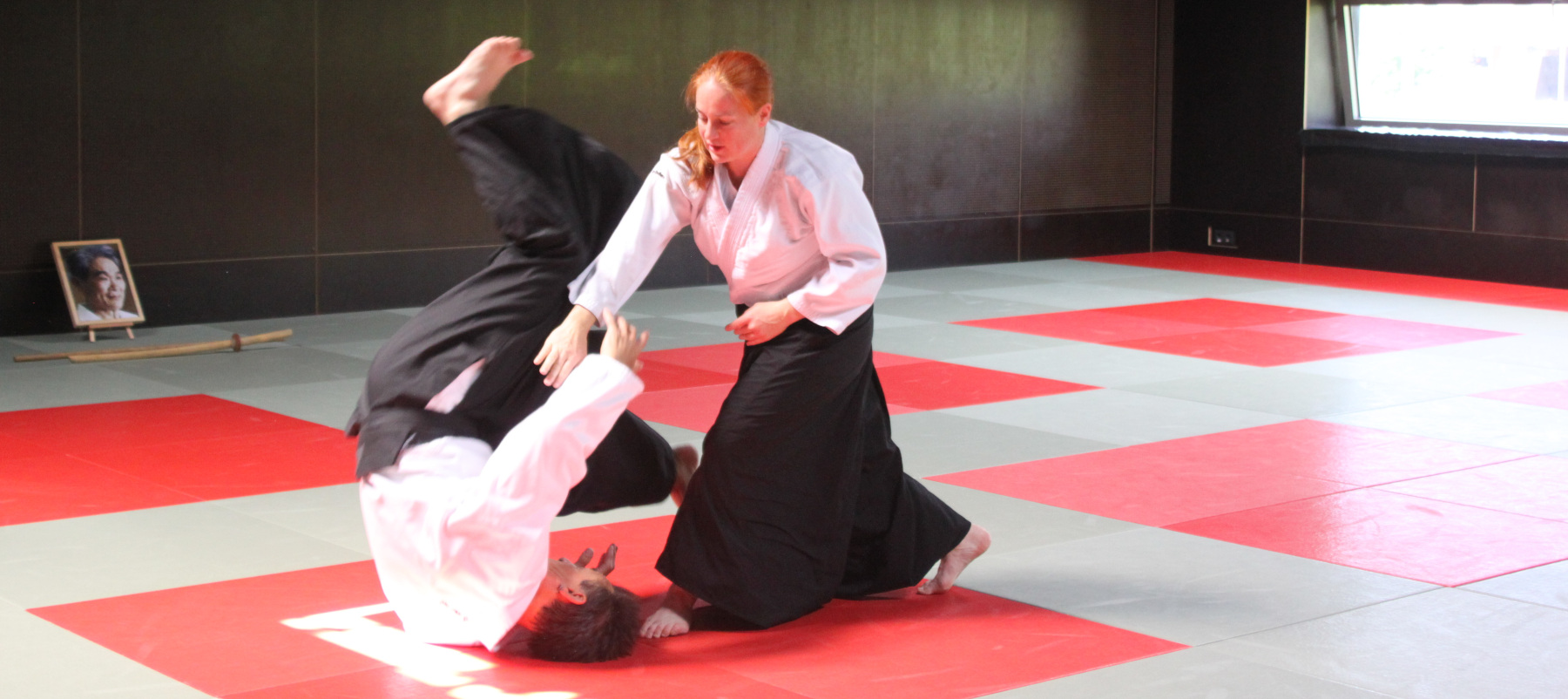 Eine Person steht breitbeinig nach vorne geneigt. Die andere Person rollt sich über den Rücken ab, um in Gegenrichtung der werfenden Person zu fliehen